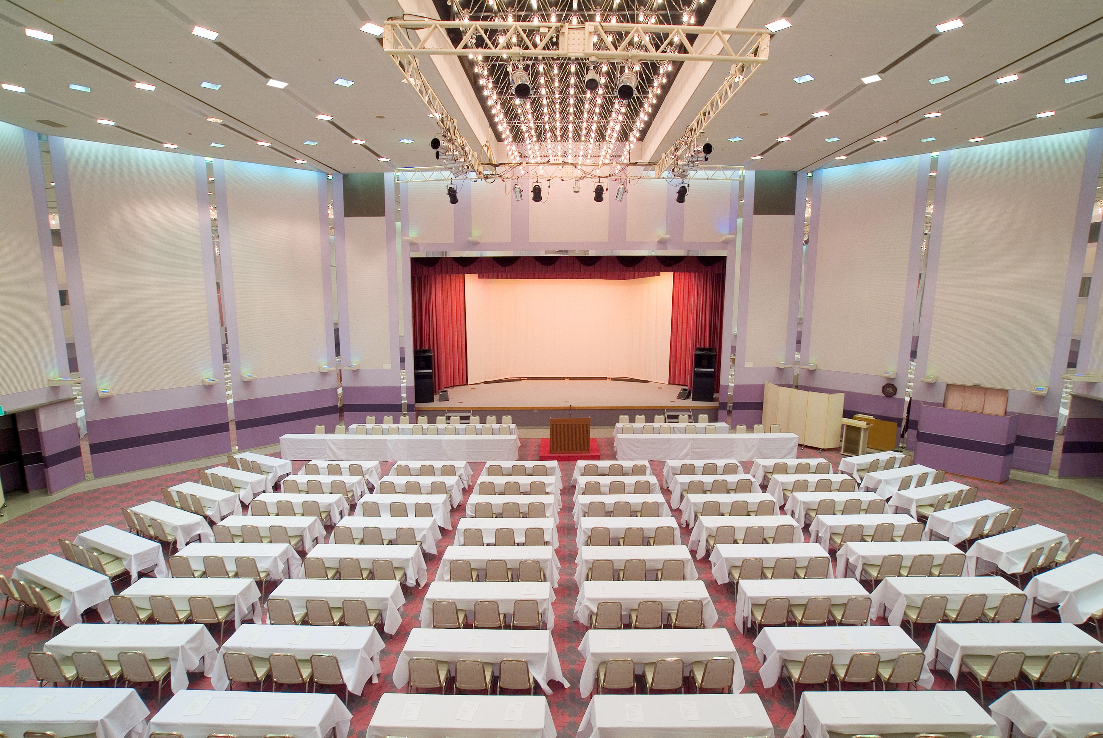 A large conference room with rows of tables and chairs, a stage with a red curtain, and a chandelier.