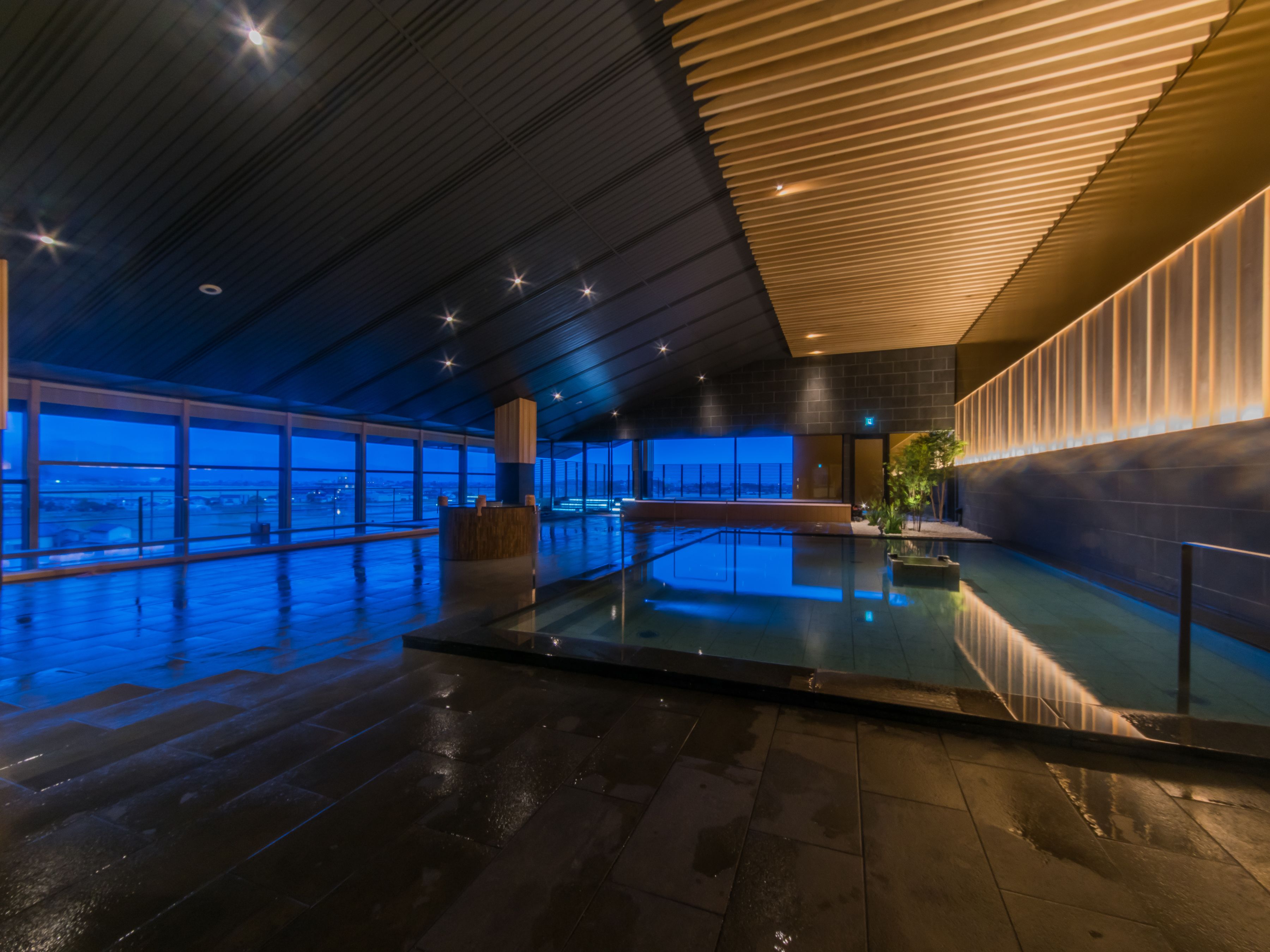 A modern indoor pool area with a glass barrier, large windows, and a wooden ceiling.