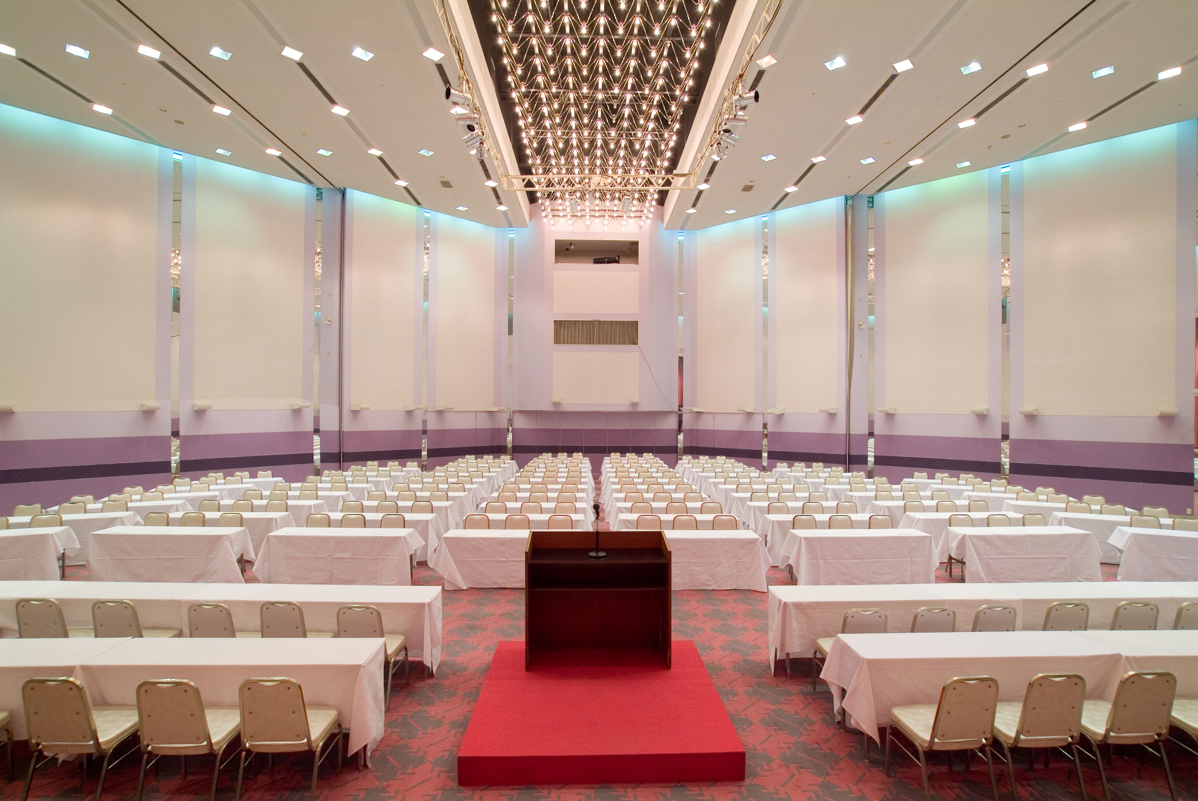 A conference room with rows of tables and chairs, a podium at the front, and a ceiling with recessed lighting.