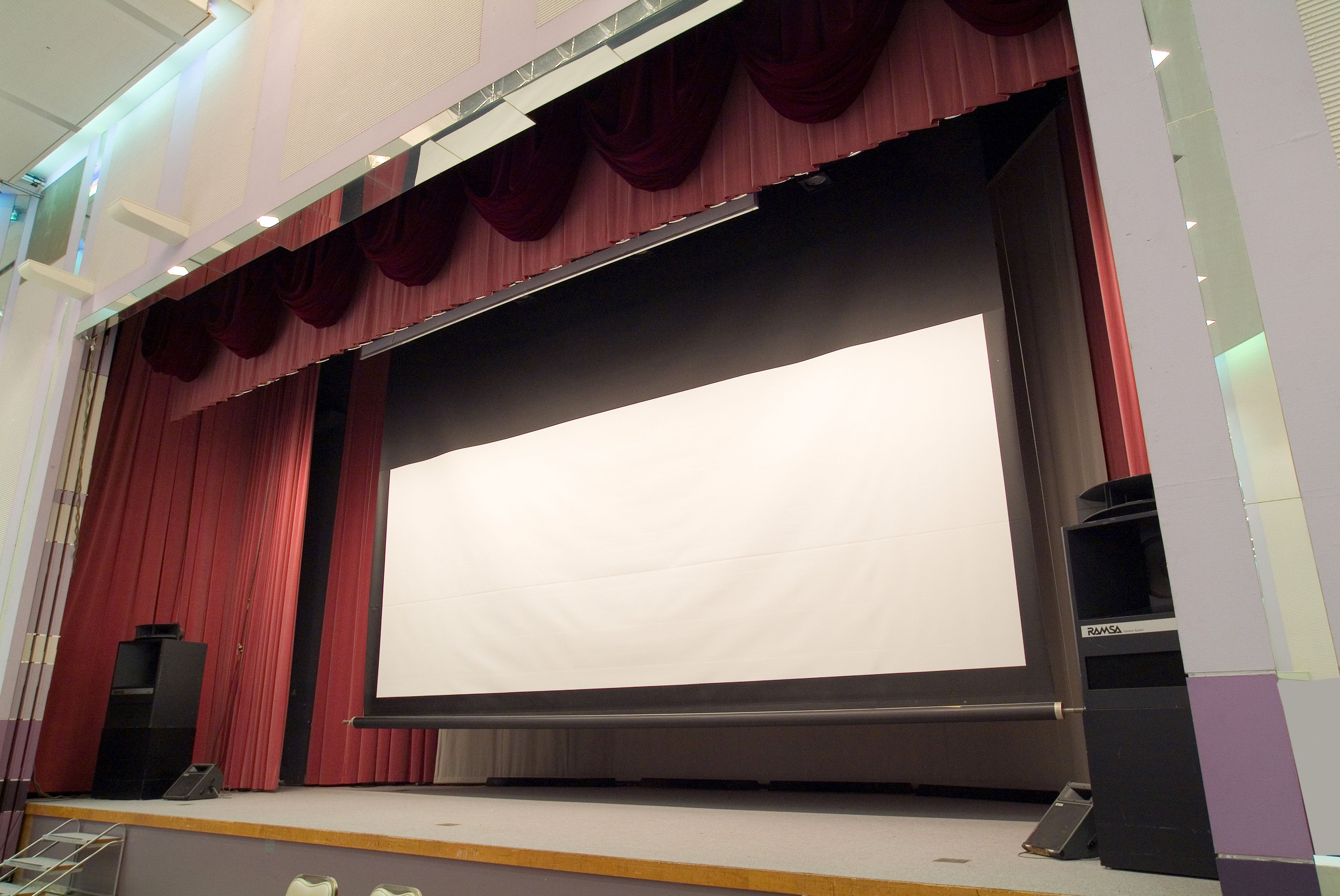 Un escenario de teatro con una gran pantalla blanca, cortinas rojas y un altavoz a la izquierda.