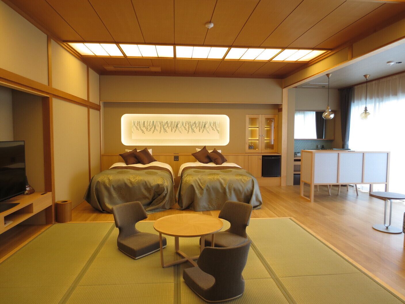 A hotel room with two beds, a TV, a small kitchen area, and a seating area with a table and chairs.