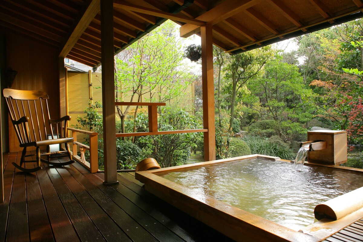 A wooden deck with a hot tub and a rocking chair, surrounded by lush greenery.