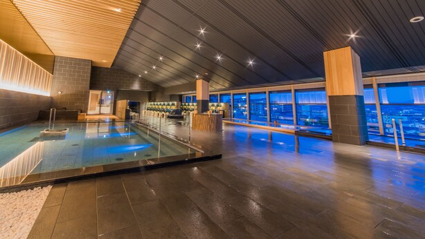 A modern indoor pool area with a glass barrier, a bar, and a view of the city at night. - Grandia Housen (Awara)