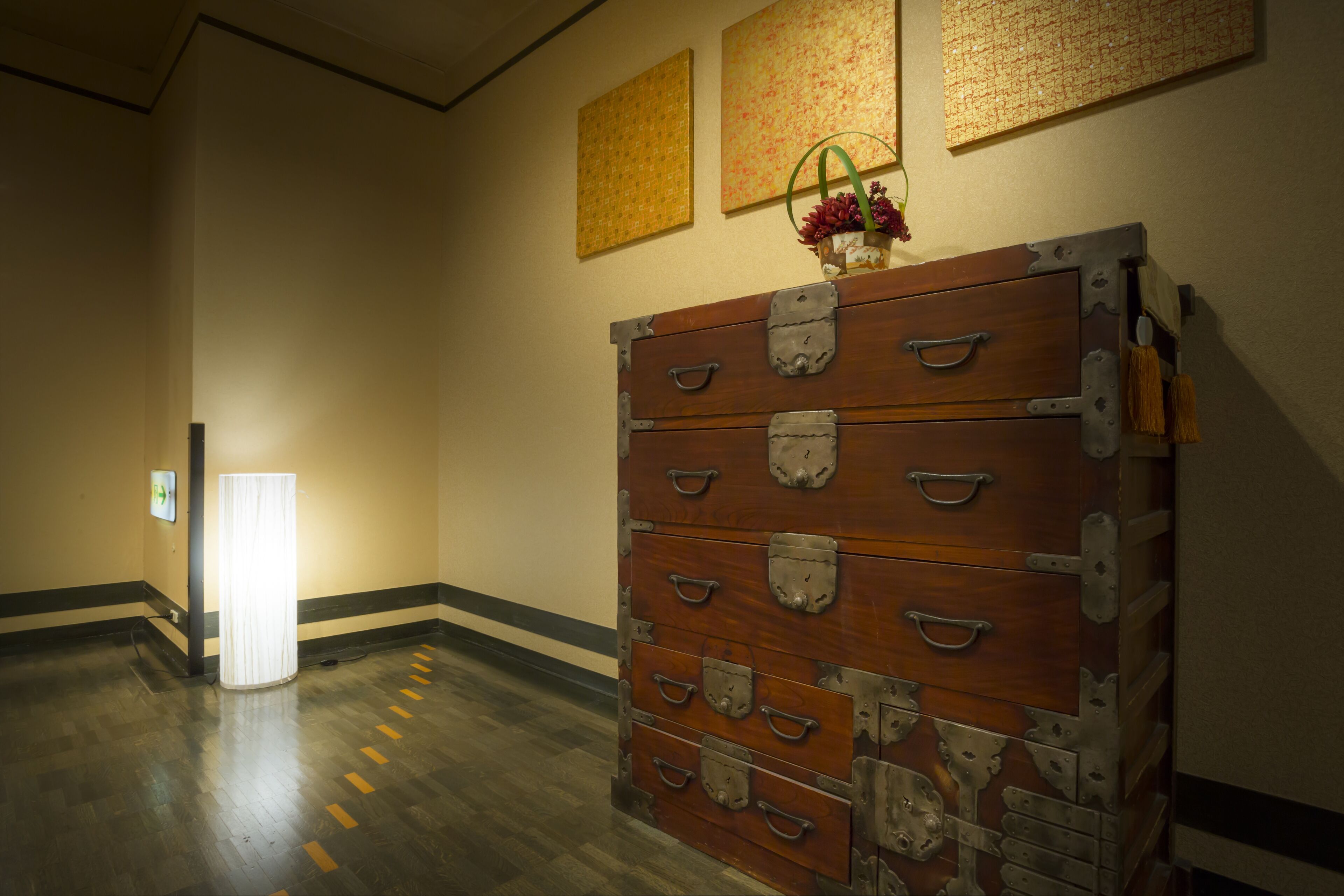 A hallway with a large wooden chest with multiple drawers, a light source on the left, and three framed artworks on the wall.