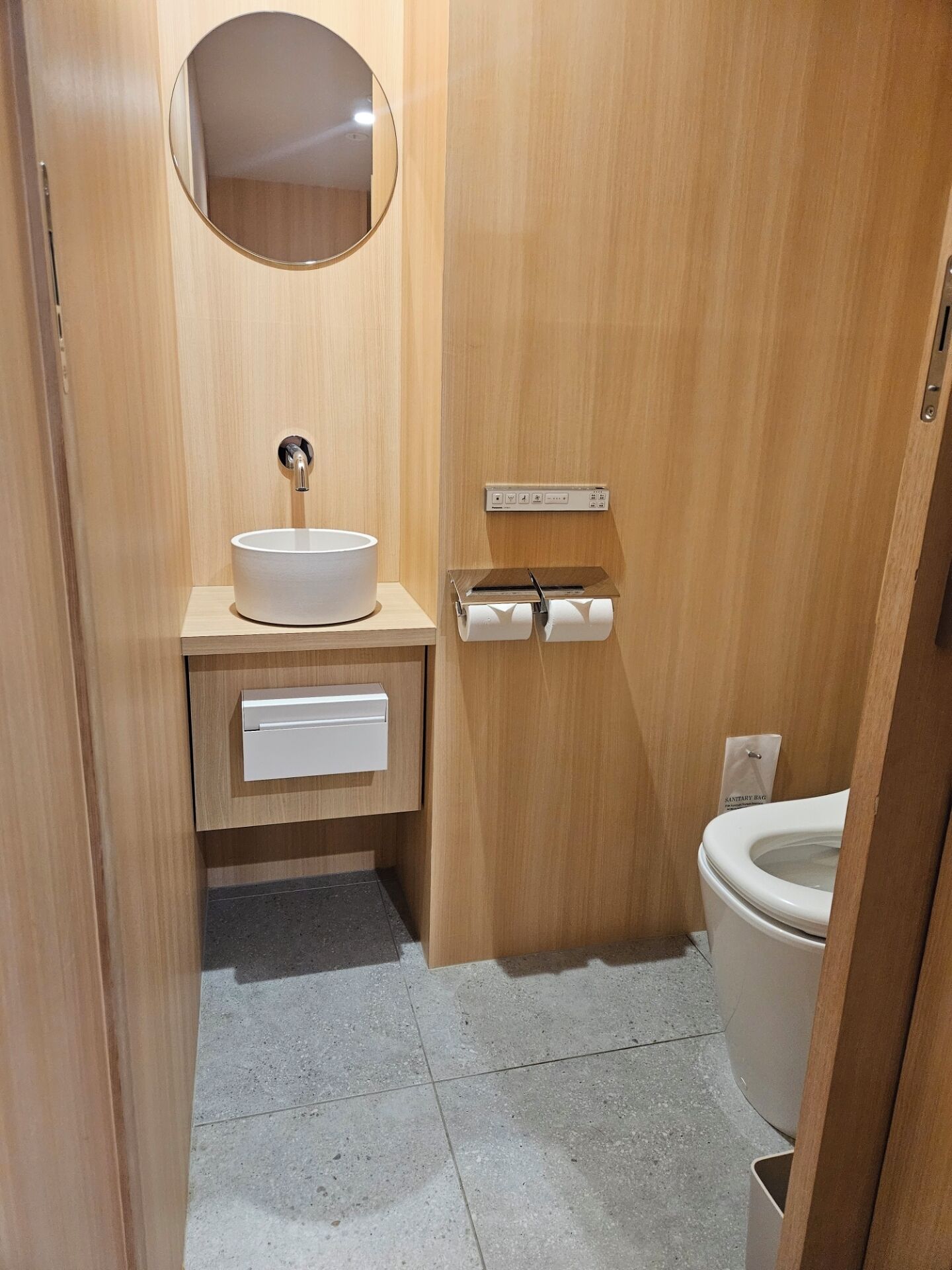 A compact bathroom with a wooden vanity, a round mirror, a toilet, and a paper towel dispenser.