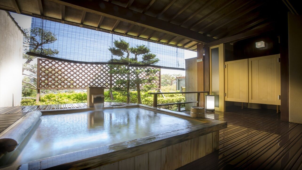 A hot spring bath area with wooden flooring, a wooden bench, and a view of greenery through a lattice screen.
