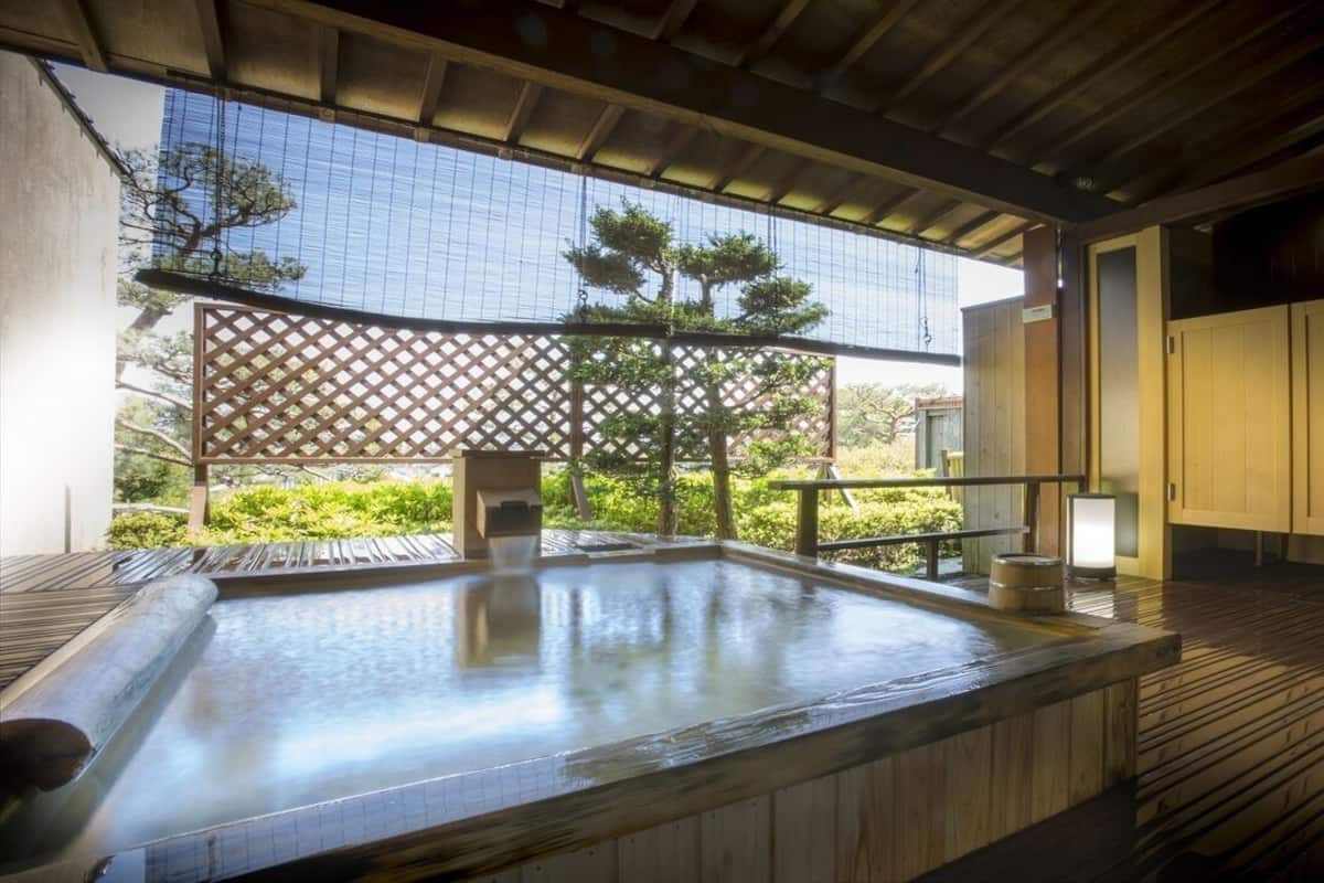 Zona de baño termal con suelo de madera, un banco de madera y vistas a la vegetación a través de una celosía.