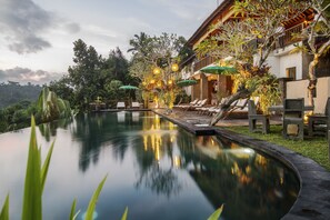 Une piscine à débordement paisible, offrant une vue dégagée sur la végétation luxuriante environnante, ainsi qu’un bâtiment doté d’un espace extérieur avec des sièges et des parasols.