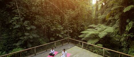A wooden deck with two individuals meditating on pink mats, surrounded by dense forest.