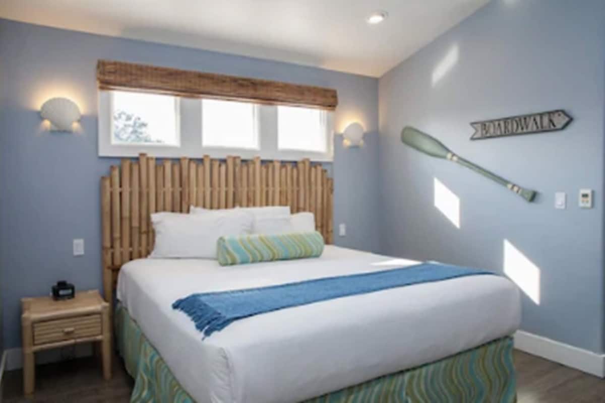 A bedroom with a bed, a nightstand, a window with blinds, and a wall-mounted sign.