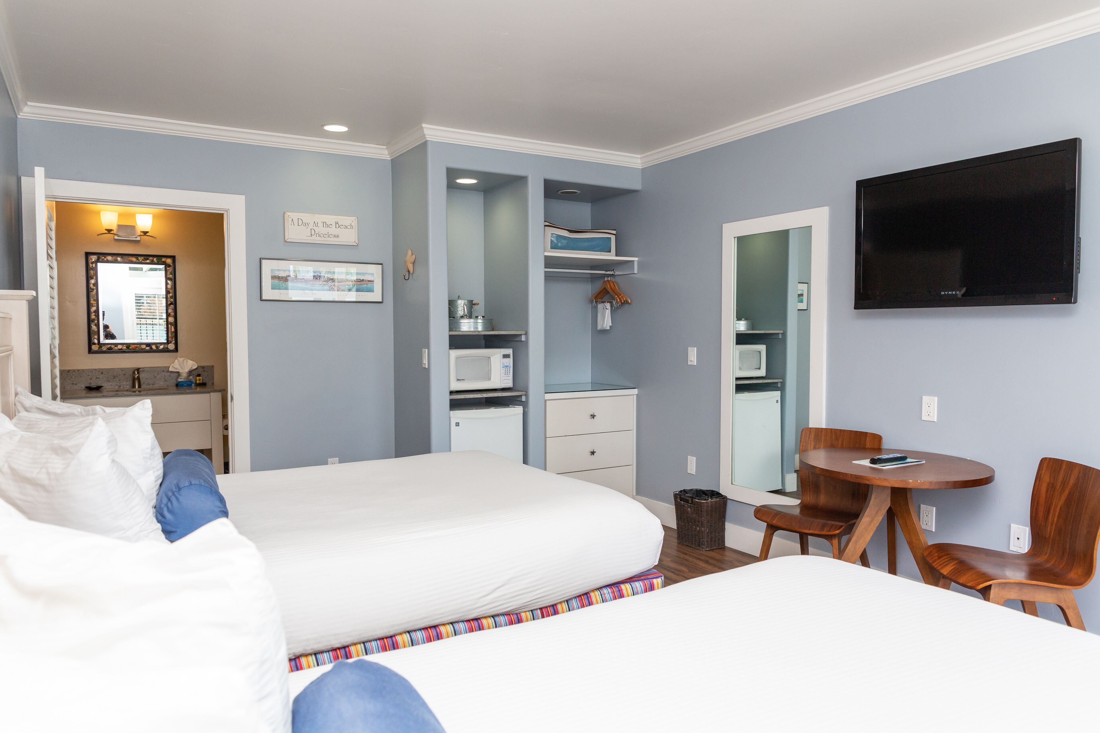 A hotel room with two beds, a desk with a chair, a TV, and a small kitchenette.