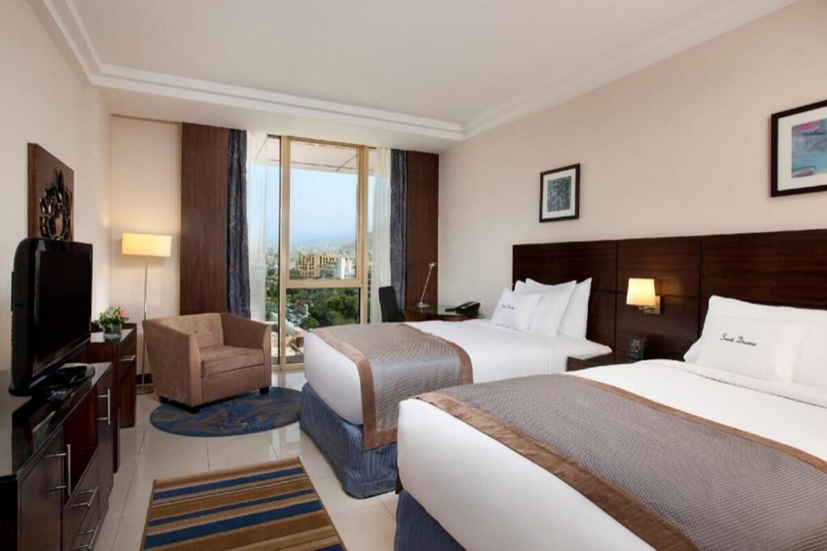 A hotel room with two beds, a TV, a desk, a chair, and a view of the city.