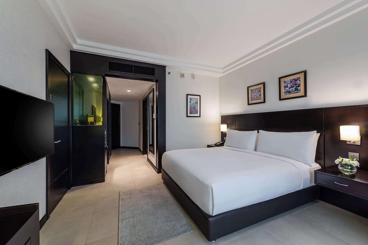 A modern hotel room with a large bed, a flat-screen TV, a desk with a lamp, and a door leading to a bathroom.