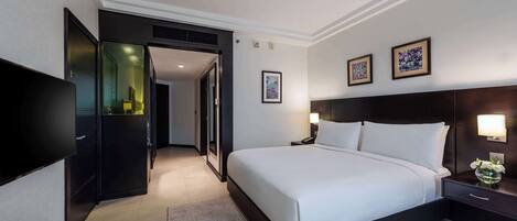 A modern hotel room with a large bed, a flat-screen TV, a desk with a lamp, and a door leading to a bathroom.