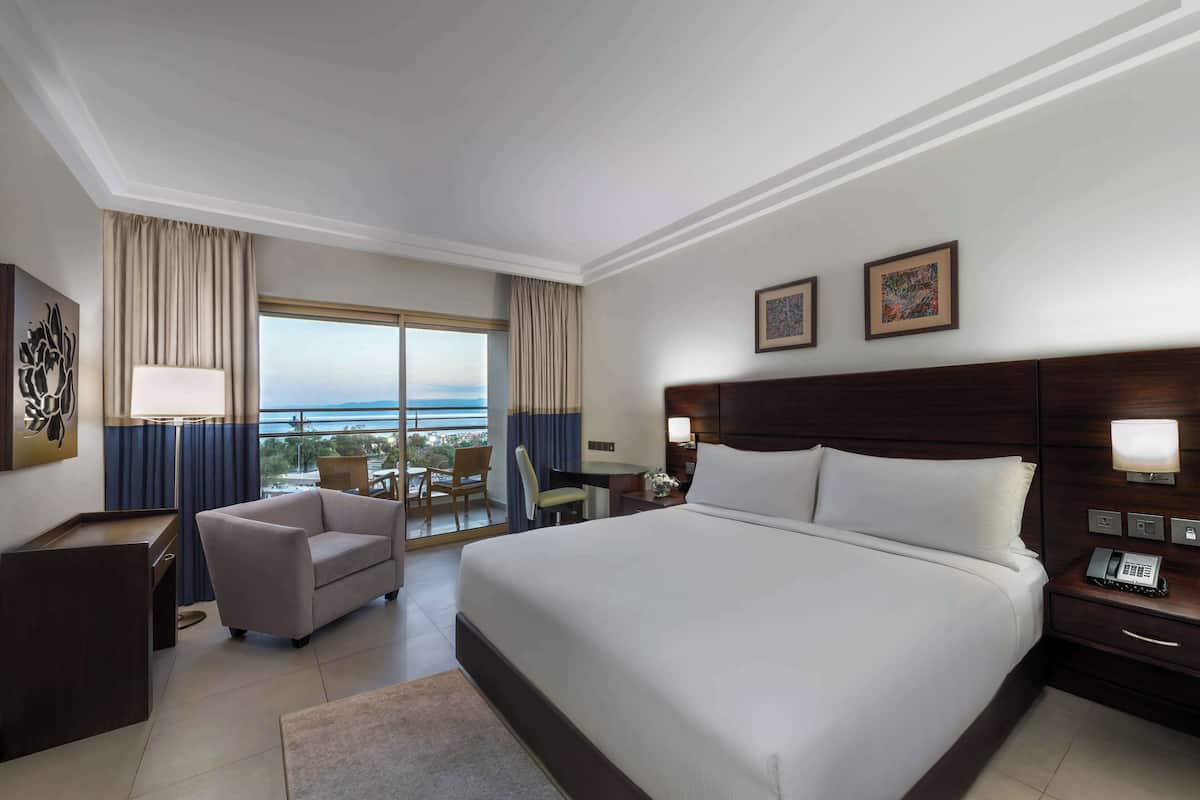 A hotel room with a large bed, a desk, a chair, a desk lamp, a phone, and a view of the outdoors.