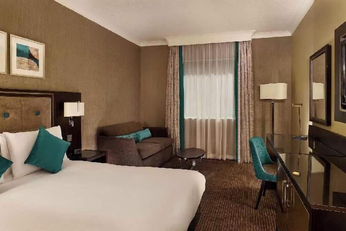 A hotel room with a bed, a desk, a chair, a sofa, a TV, and two framed pictures on the wall.