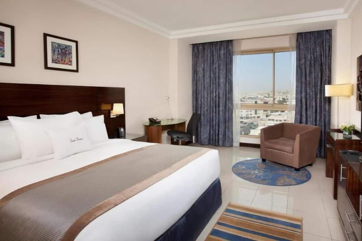 A hotel room with a bed, a desk, a chair, a view of the city, and a large window with curtains.