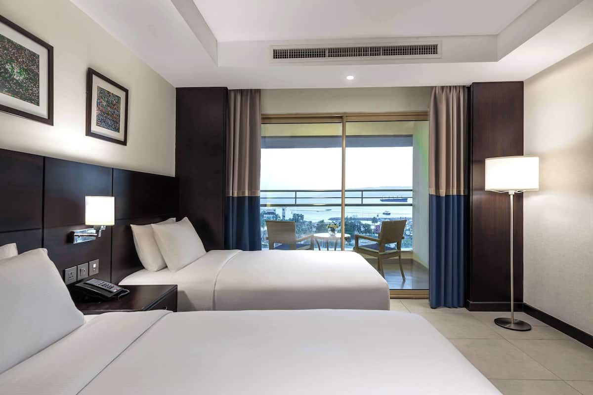 A hotel room with two beds, a desk with a phone, a lamp, and a view of the city through large windows.