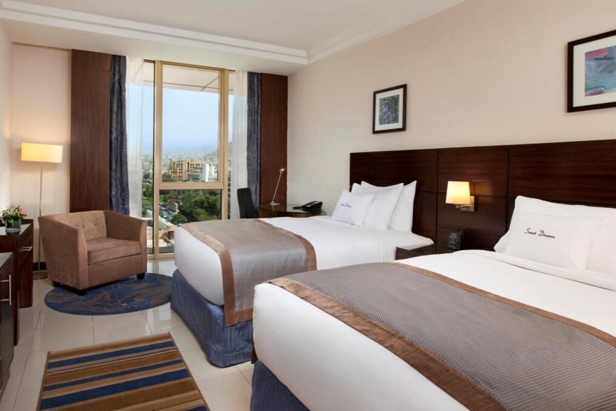 A hotel room with two beds, a desk, a chair, a window with a city view, and a framed picture on the wall.