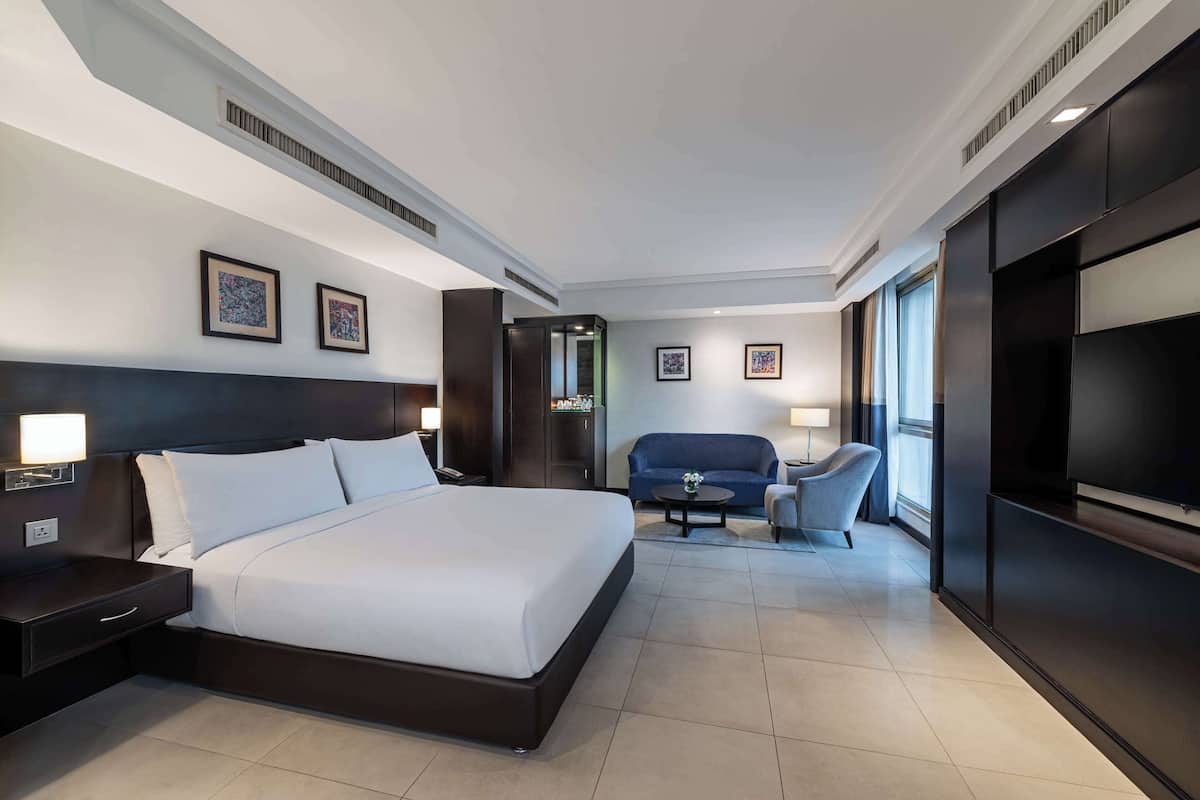 A modern hotel room with a large bed, a seating area with a sofa and armchairs, a flat-screen TV, and artwork on the walls.