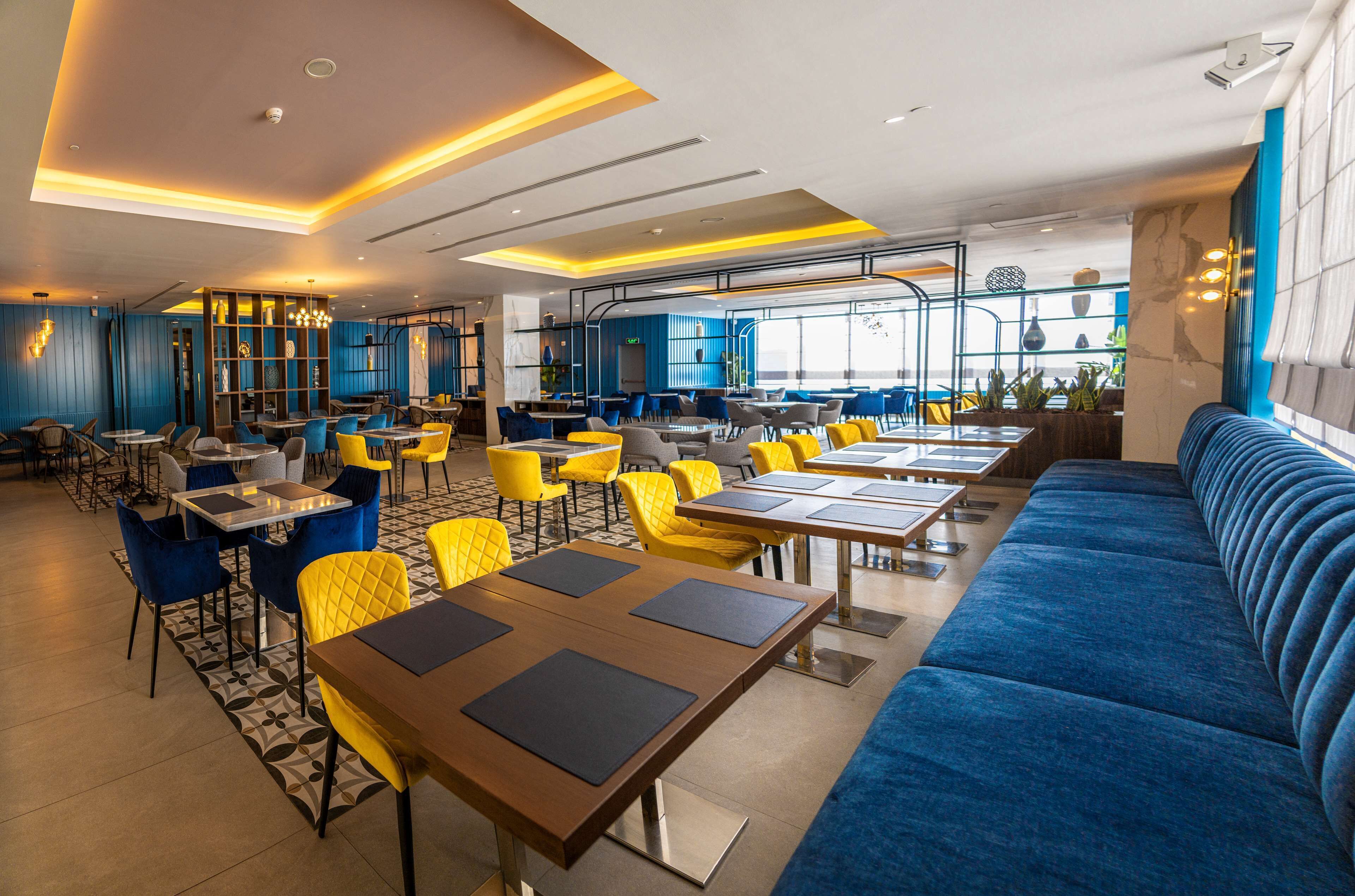 Un restaurant modern amb seients blaus i grocs, finestres grans i un sofà blau.