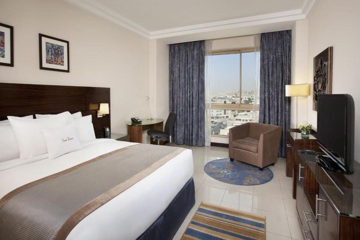 A hotel room with a large bed, a desk, a chair, a television, and a view of the city.