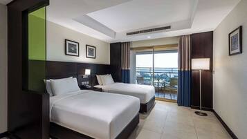 Habitación de hotel con dos camas, un ventanal con cortinas, una lámpara de pie y vistas a la ciudad.