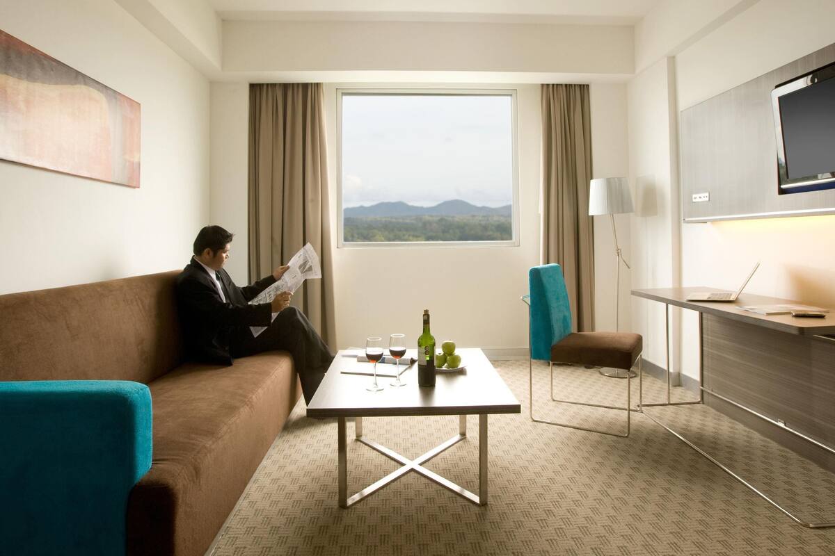 A hotel room with a large window, a sofa, a small table with a bottle and glasses, a chair, and a desk with a laptop.