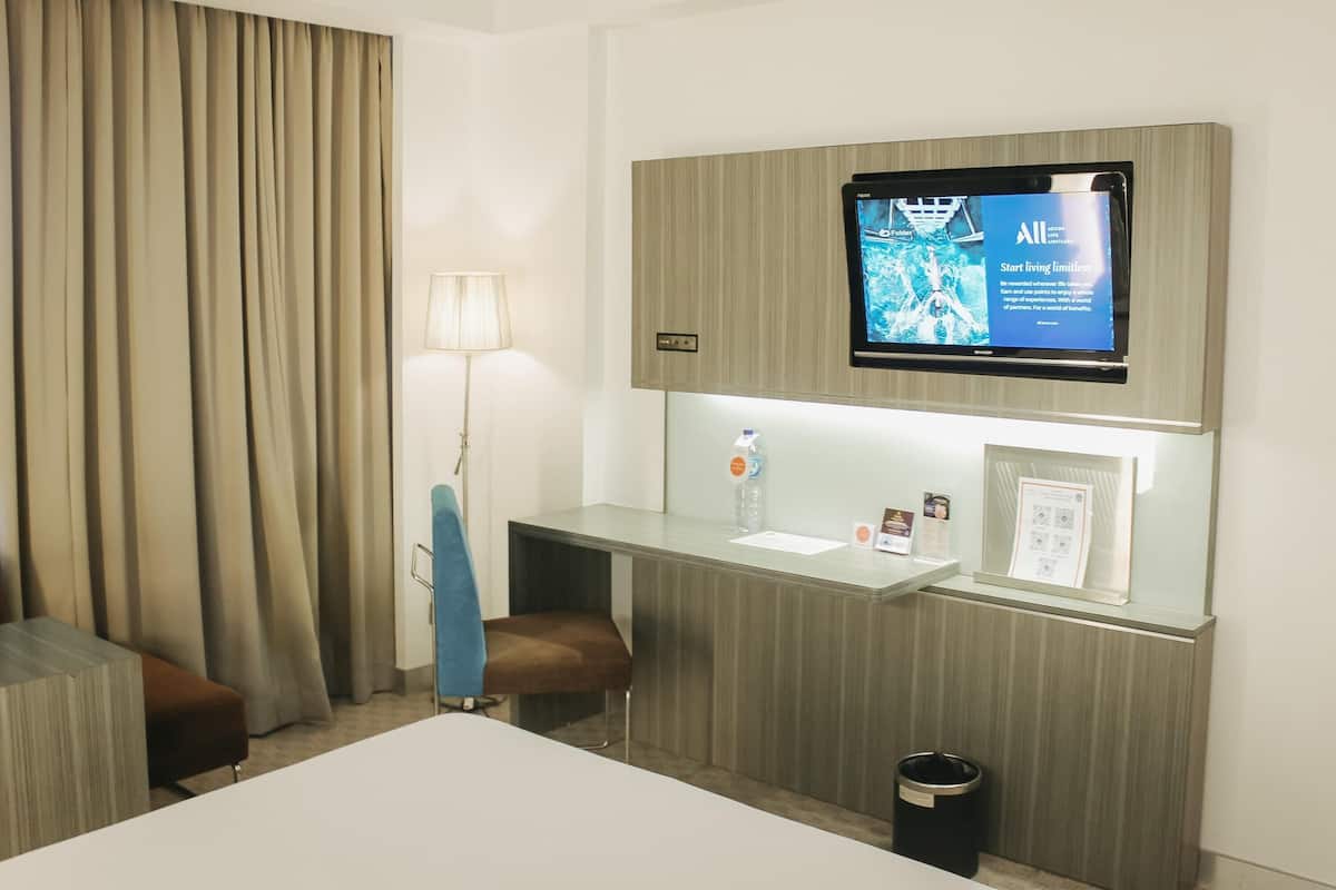 A hotel room with a bed, a desk with a chair, a television mounted on the wall, a lamp, and a trash can.