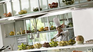 A chef preparing food in a well-lit kitchen with various fruits and vegetables displayed on shelves and counters.