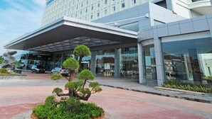 A modern hotel with a large glass facade, a covered entrance, and a landscaped area with a central tree.
