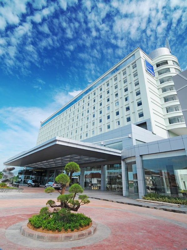 A modern hotel with a large glass facade, a covered entrance, and a landscaped area with a central tree.