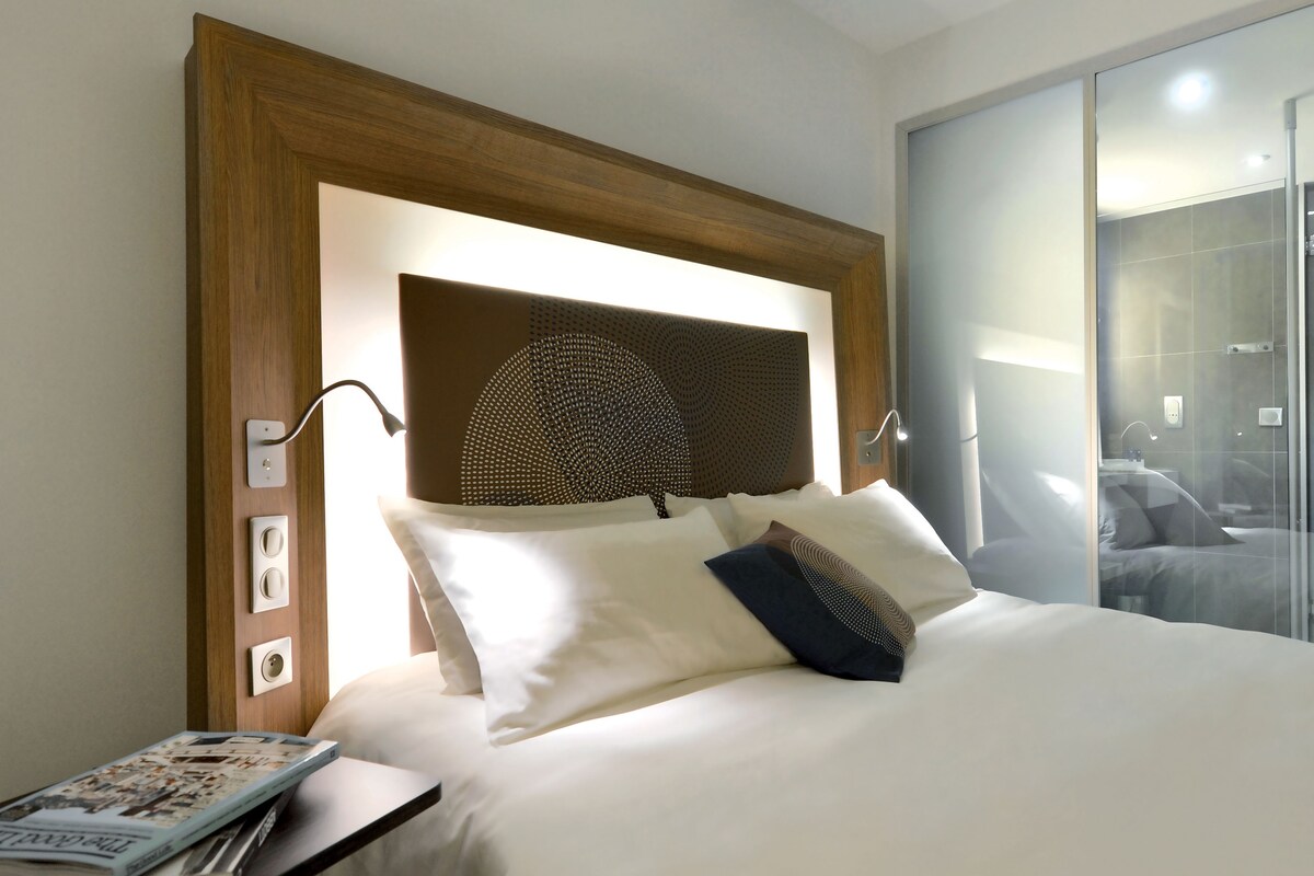 A hotel room with a large bed, a wooden headboard, a bedside table with a magazine, and a glass shower door.