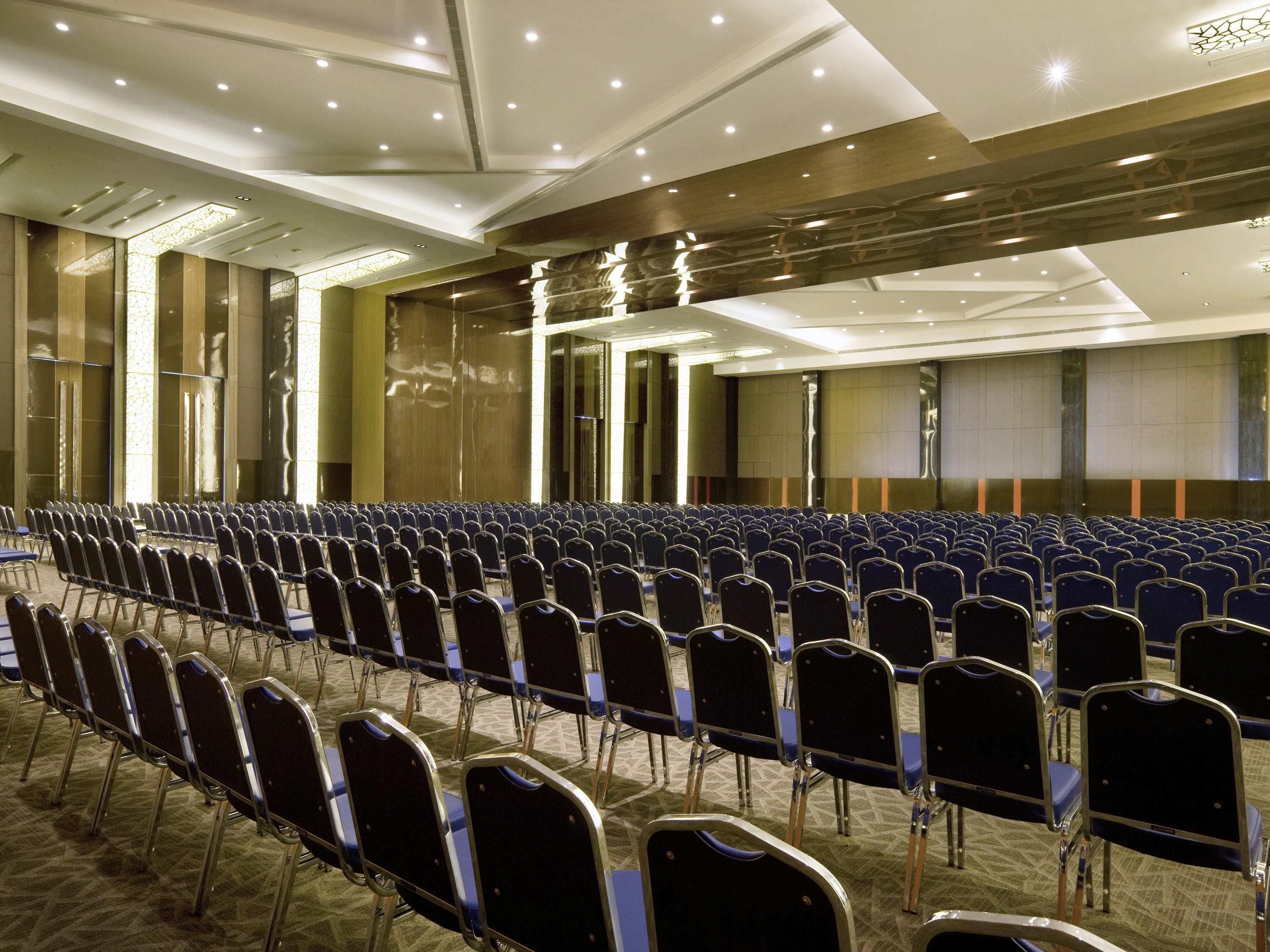 Une salle de conférence avec des rangées de chaises, un sol recouvert de moquette et un plafond haut doté d’un éclairage encastré.