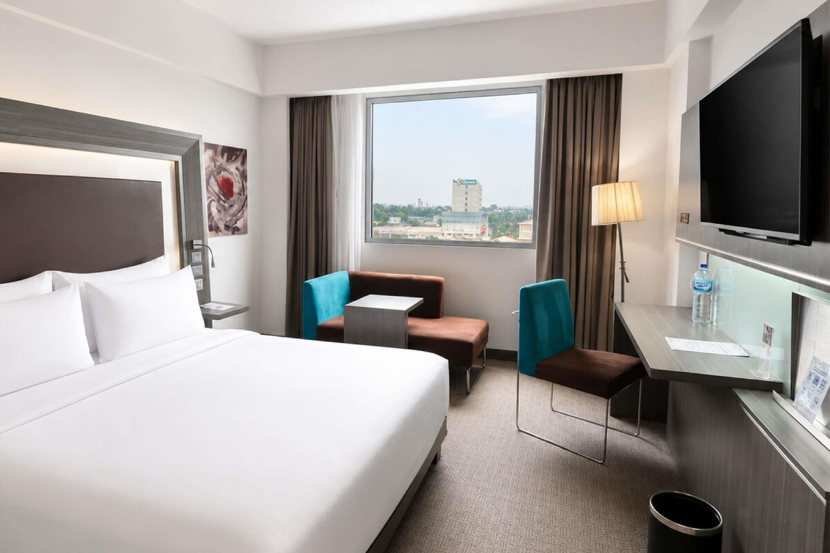 A hotel room with a bed, a desk, a chair, a TV, and a window with a view of the city.