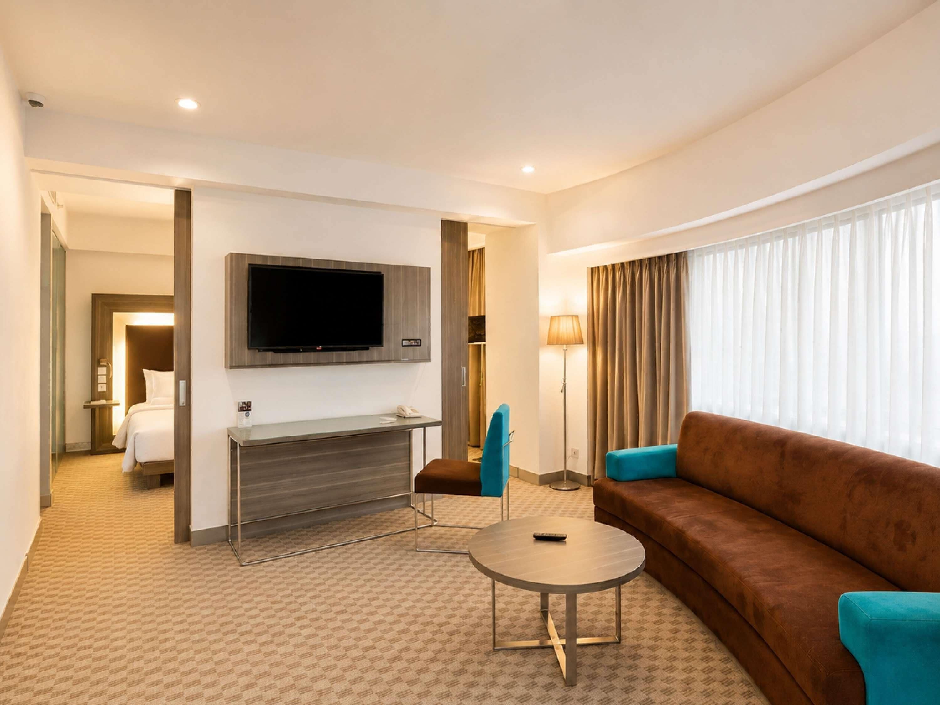 Quarto de hotel com televisão de tela plana na parede, um sofá marrom, uma mesa de centro redonda e uma poltrona azul.