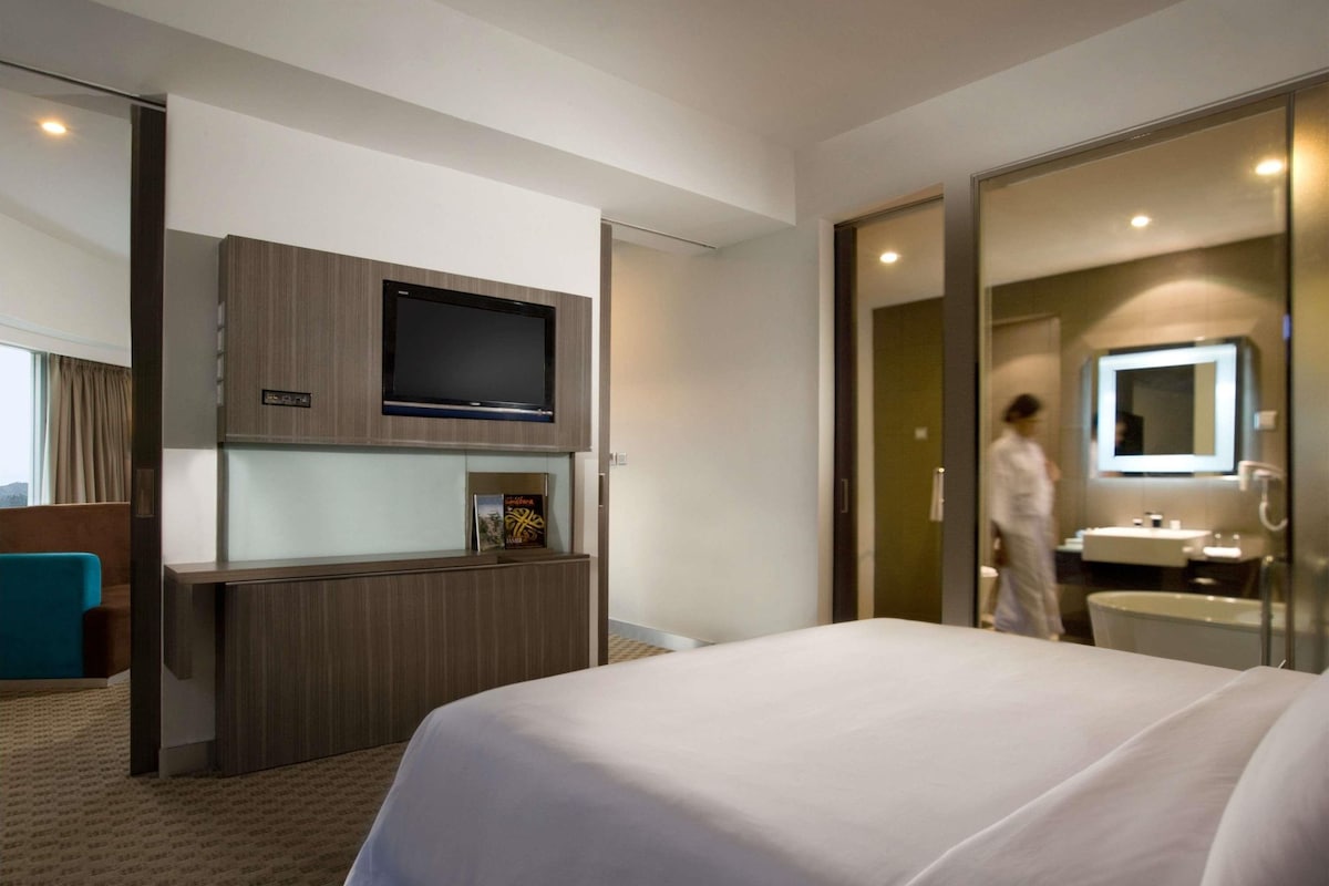 A hotel room with a bed, a TV mounted on a wooden panel, a bathroom with a sink and mirror, and a person walking in the background.