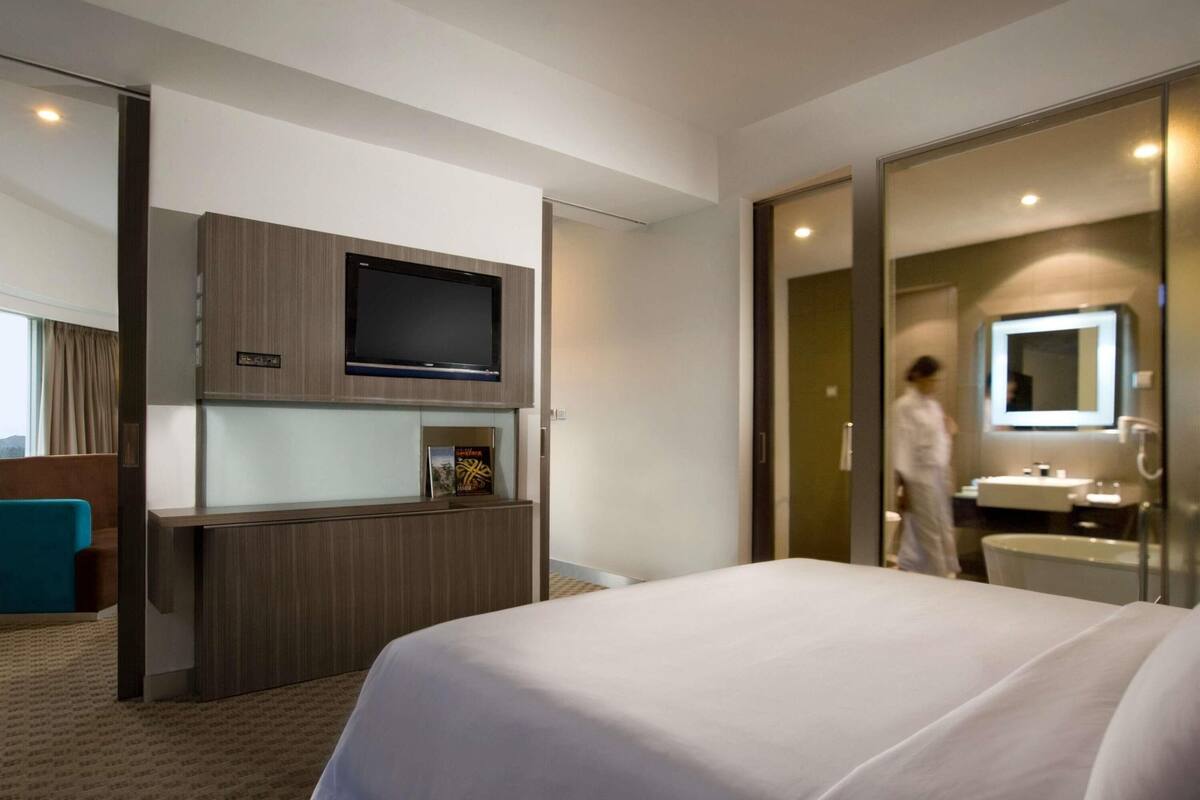 A hotel room with a bed, a TV mounted on a wooden panel, a bathroom with a sink and mirror, and a person walking in the background.