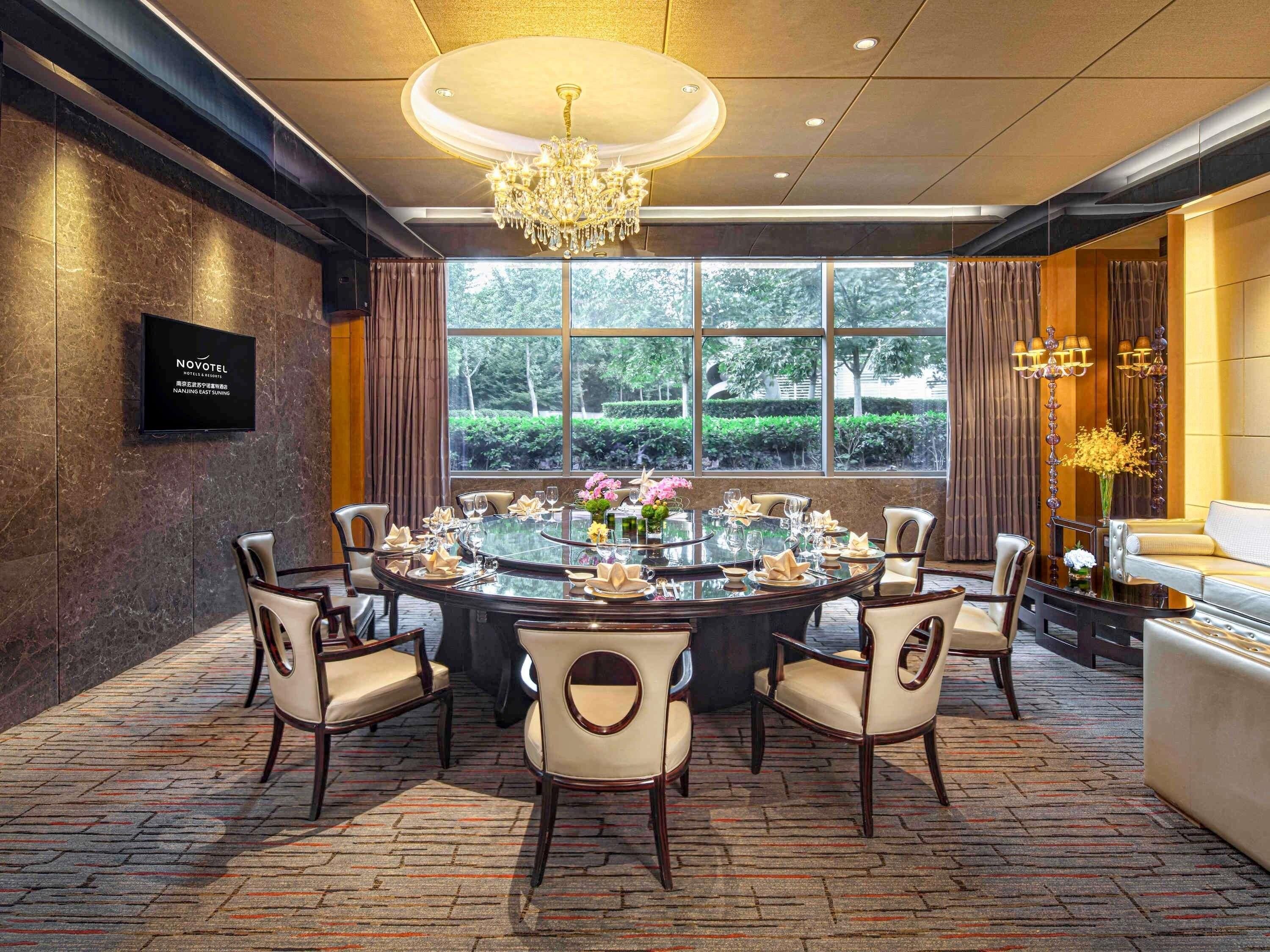 A hotel dining room with a round table, chairs, a chandelier, and a view of greenery outside.