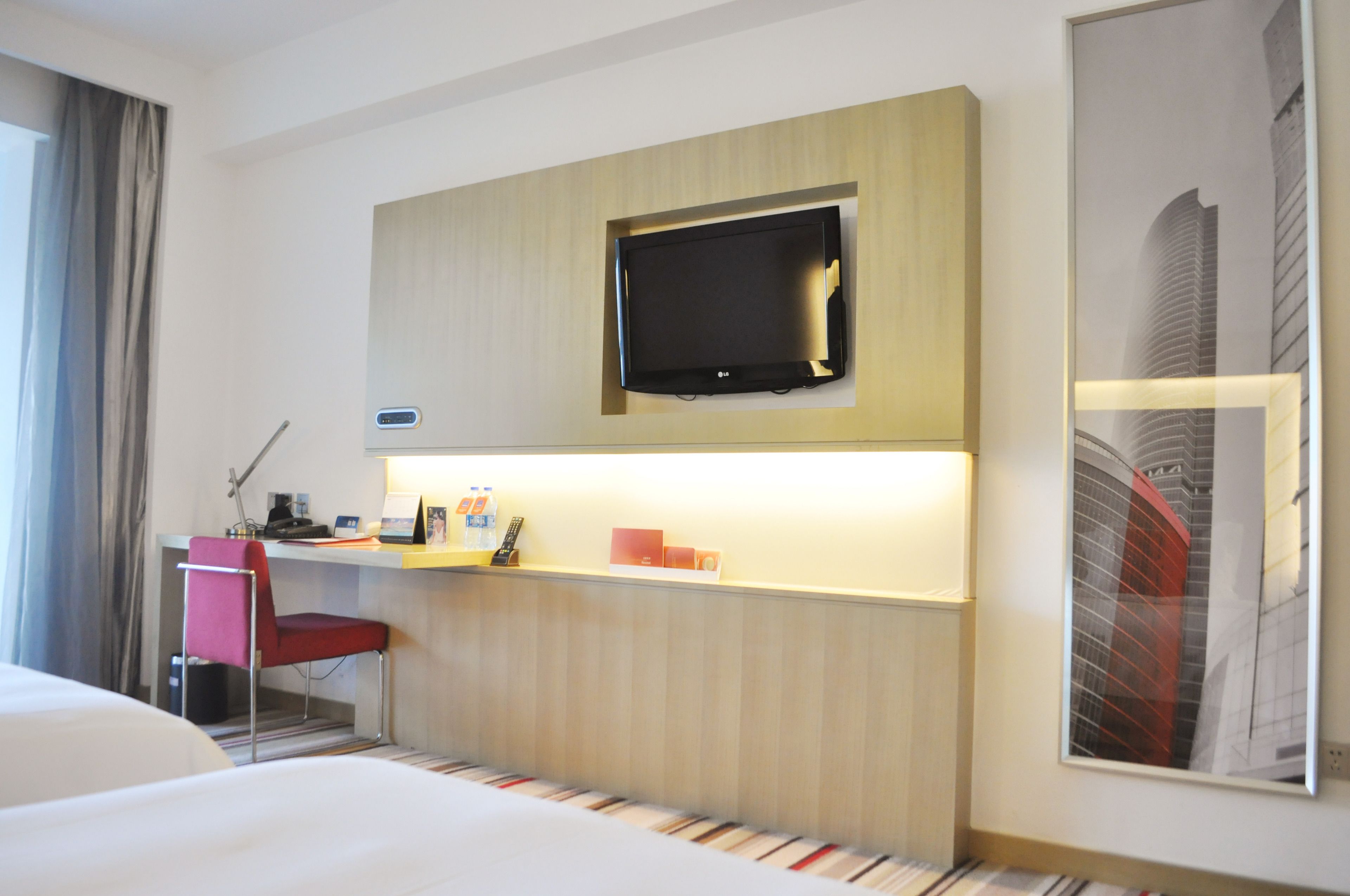 Habitación de hotel con cama, escritorio con silla, televisor de pantalla plana montado en la pared y un cuadro enmarcado.