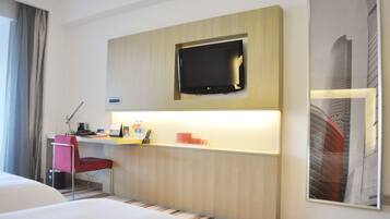 Habitación de hotel con cama, escritorio con silla, televisor de pantalla plana montado en la pared y un cuadro enmarcado.