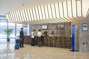 A hotel reception area with a marble reception desk, a blue pillar with the hotel name, and a large window providing natural light. - Novotel Nanjing East Suning (Nanjing)