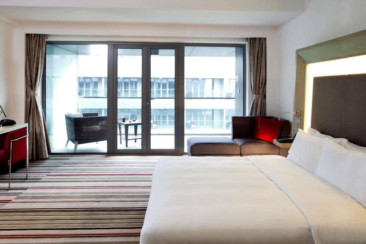Ein Hotelzimmer mit einem großen Bett, einem Schreibtisch, einem Stuhl, einem kleinen Tisch und Blick auf ein Gebäude durch eine Glastür.