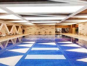 A large indoor swimming pool with a geometric floor pattern, sunlit ceiling, and lounge chairs along the sides.