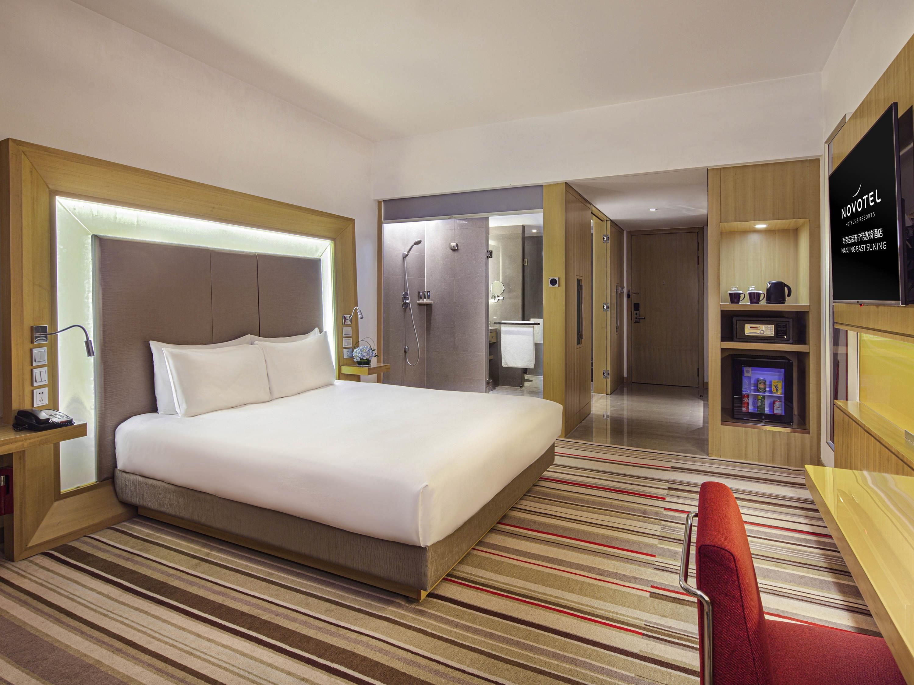 A hotel room with a large bed, a shower, a vanity with a mirror, a TV, and a minibar.