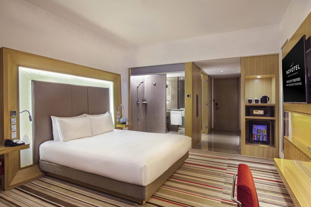 Une chambre d’hôtel avec un grand lit, une douche, une coiffeuse avec un miroir, une télévision et un minibar.