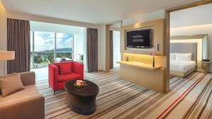 Hotelkamer met een groot raam, een flatscreen televisie, een bed, een rode stoel, een bank en een klein tafeltje met fruit.