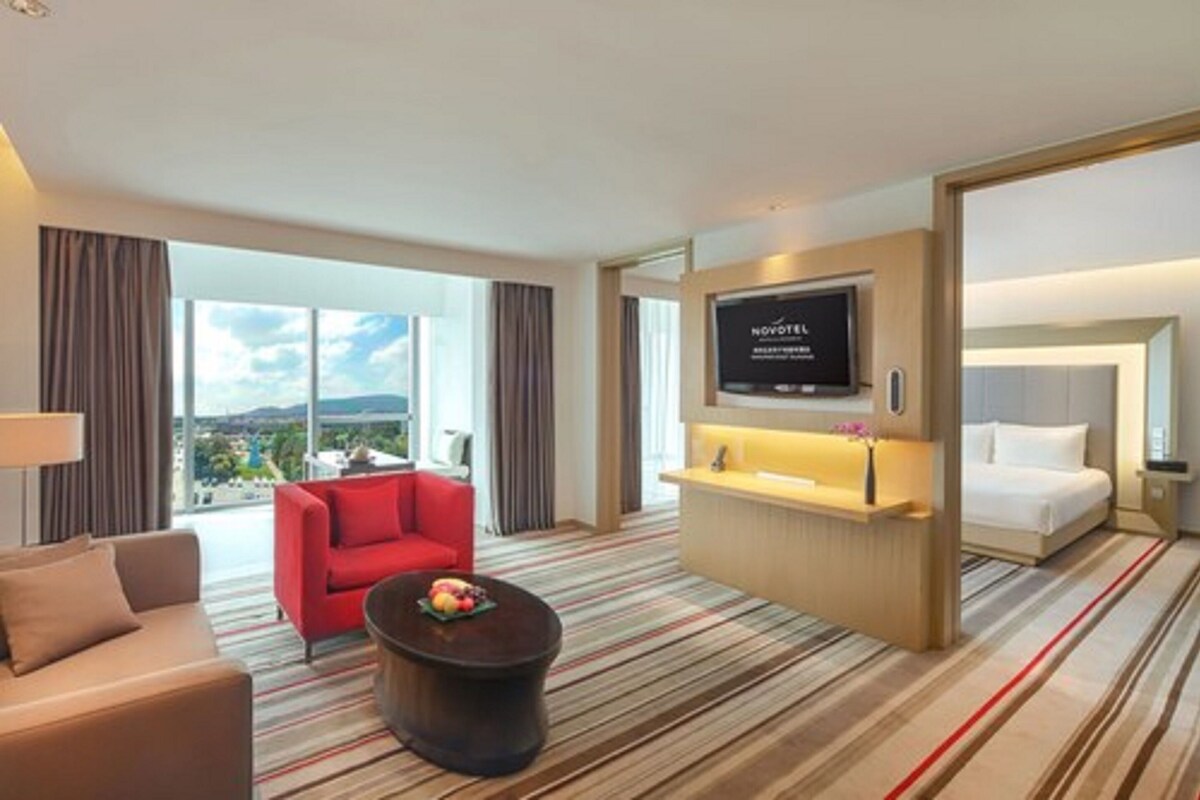 Hotelkamer met een groot raam, een flatscreen televisie, een bed, een rode stoel, een bank en een klein tafeltje met fruit.