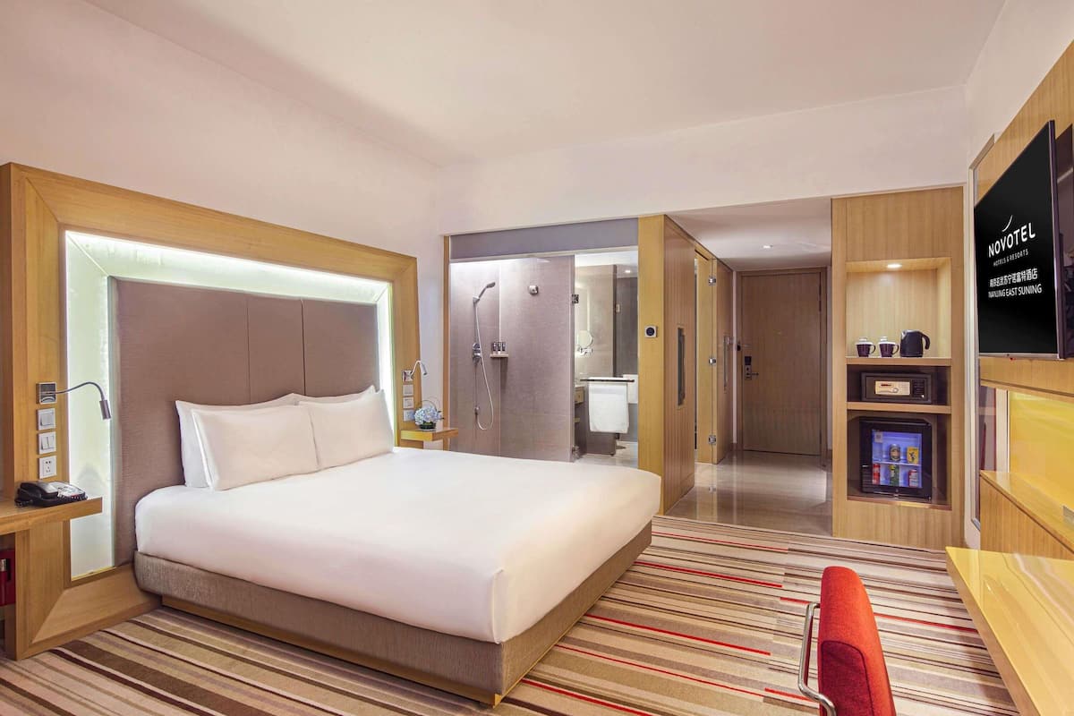 A hotel room with a bed, bedside table, nightstand, TV, minibar, and a shower area.