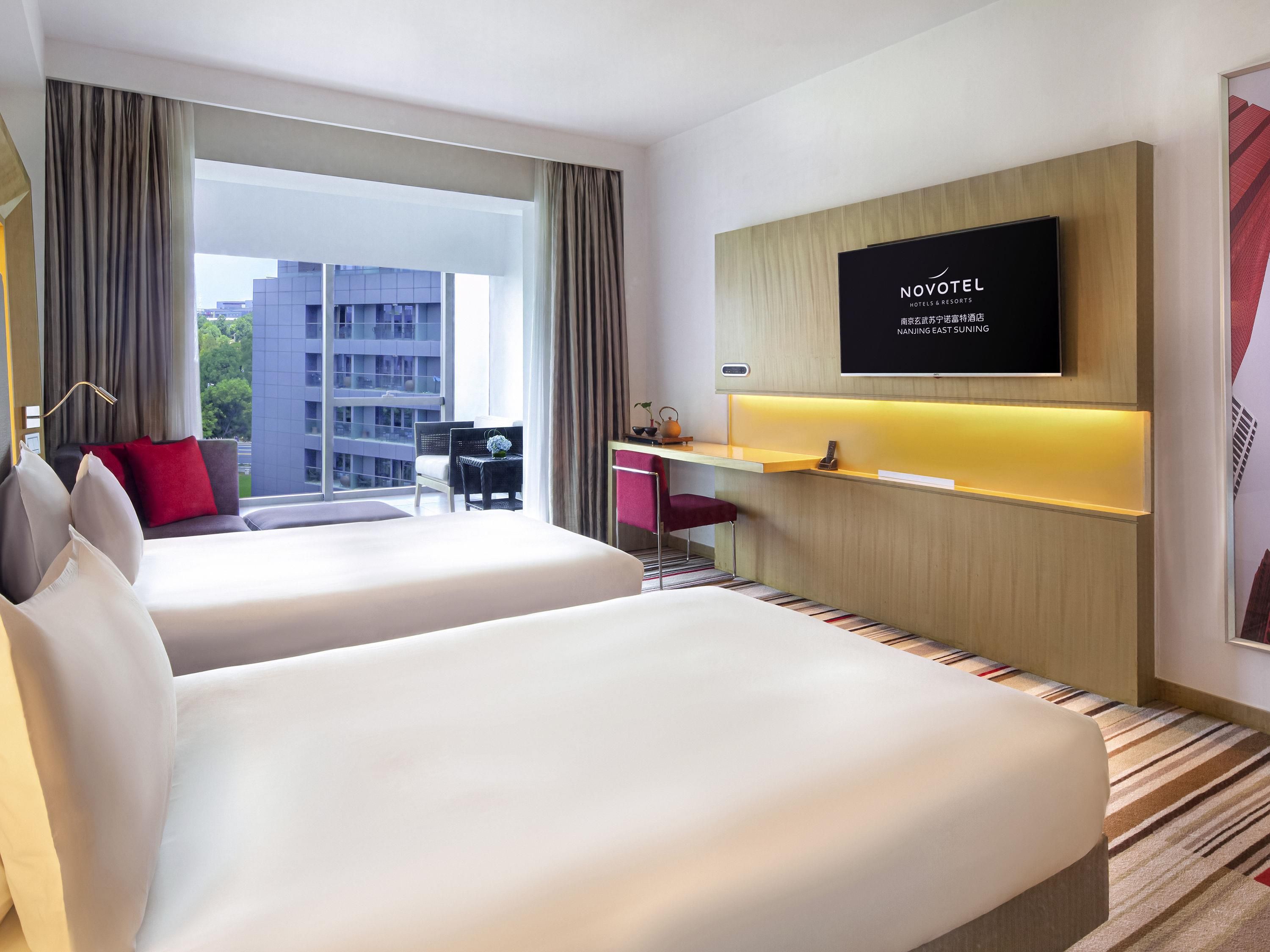 A hotel room with two beds, a desk, a TV, and a view of a building.